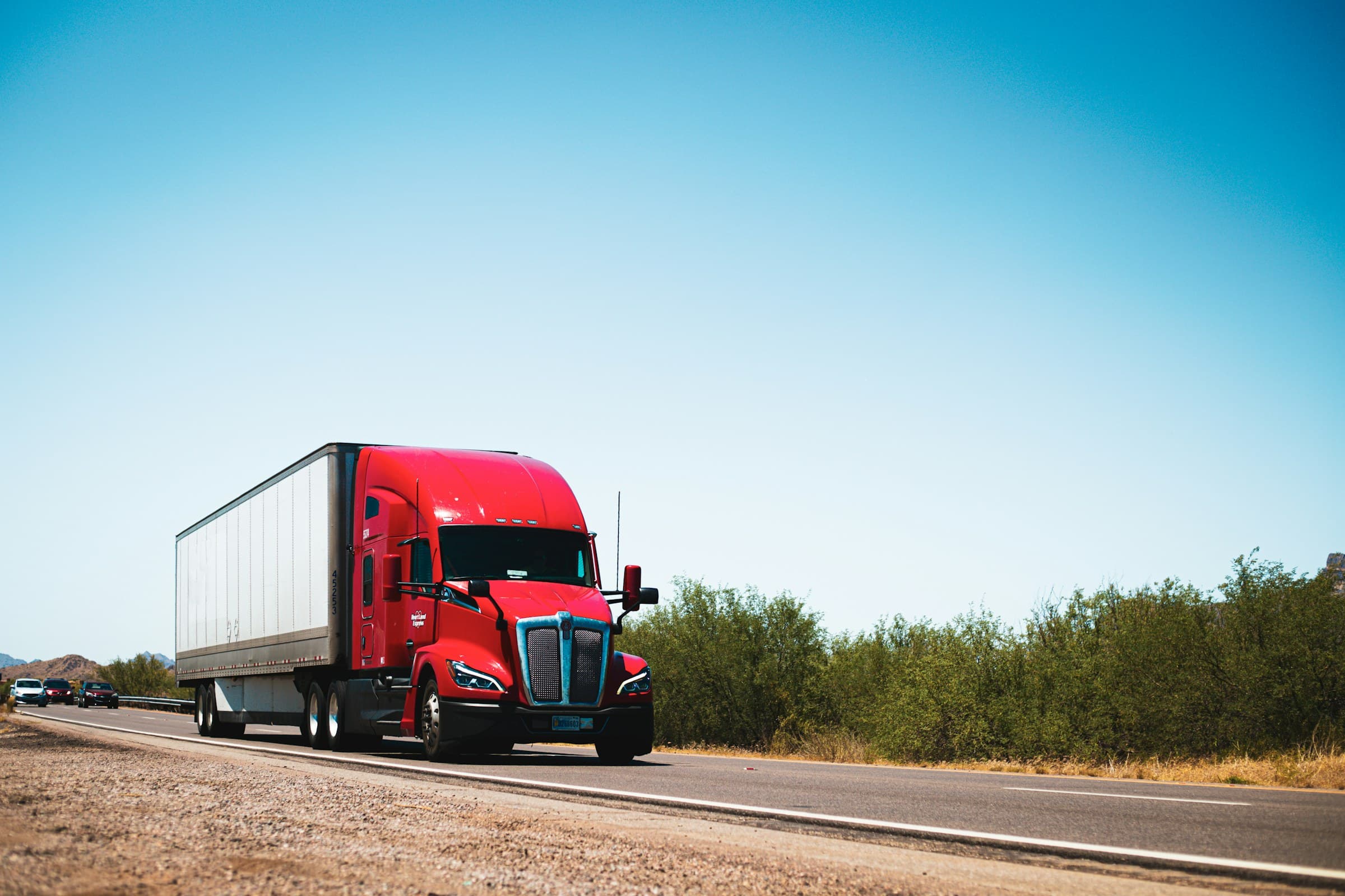 A red semi-truck hauling a dry van trailer along a US interstate under an open sky.